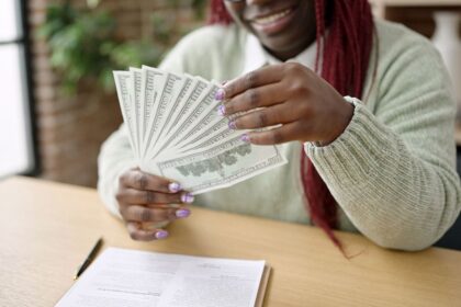 A woman sitting at her home office desk, counting cash and writing in a notebook.