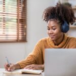 College student working from home, using a laptop, wearing headphones, and writing in a notebook.