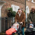 Two male friends are out in Tynemouth, North East UK. They are walking on a sidewalk and pushing their baby sons in strollers. They are wearing warm clothing.