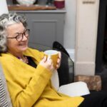 A mature adult sitting by a fireplace in a living room at home. She is wearing a yellow cardigan and spectacles.