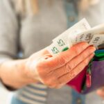 Close-up image depicting a woman in her 70s taking British bank notes from her colourful leather wallet.