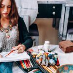 A woman sitting outdoors at a table and using colored pencils to draw in a large sketchbook.