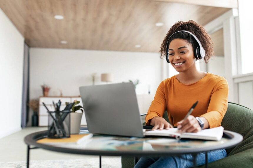A freelancer working remotely from her kitchen table, using a laptop, wearing headphones, and writing in a notebook.