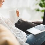 A woman working from home, drinking a cup of coffee while she uses her laptop.