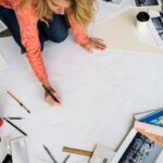 A woman working on a vision board on the floor, drawing on a large posterboard, with magazine cuttings and collage items in the background.
