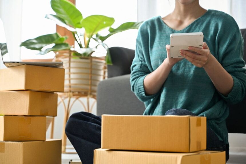 An online reseller holding a tablet and packing boxes while sitting on the living room floor.