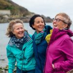 Friends and sisters exploring the outdoors together in Cornwall. They are standing with their arms around each other at the coast.
