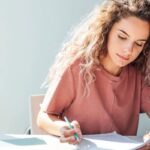 A woman sitting at a desk, working on a laptop and writing in a notebook.