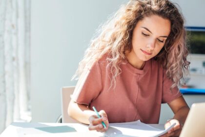 A woman sitting at a desk, working on a laptop and writing in a notebook.