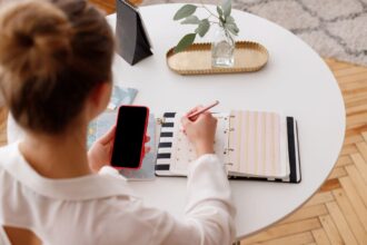 Overhead view of a woman using a smartphone and writing in a notebook at a desk.
