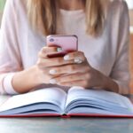 A woman using a smartphone while sitting at a table and reading a book.