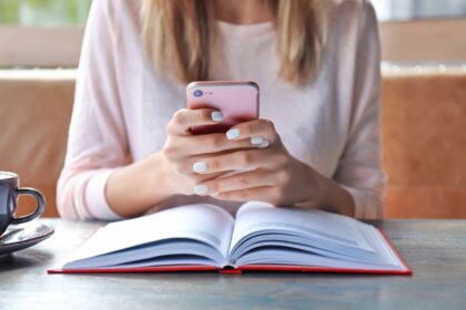 A woman using a smartphone while sitting at a table and reading a book.