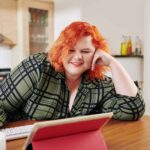 Happy young plus size woman sitting at kitchen table and watching tv series on tablet computer
