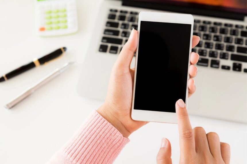 A closeup image of a woman's hands using a smartphone while sitting in front of a computer.