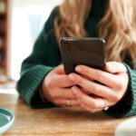 A woman sitting at a table, drinking coffee, and using a smartphone.