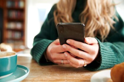 A woman sitting at a table, drinking coffee, and using a smartphone.