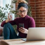 Woman sitting at her home office desk, using her phone while drinking coffee.