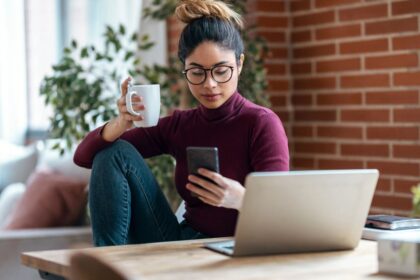Woman sitting at her home office desk, using her phone while drinking coffee.