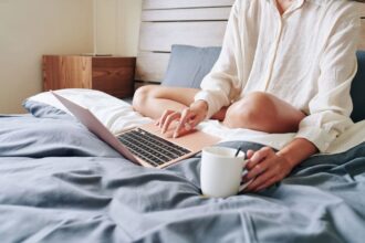 A woman working from home, using her laptop in bed while wearing pajamas.