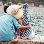 A couple sitting together on a rock wall, overlooking a lake with hills and buildings in the distance.
