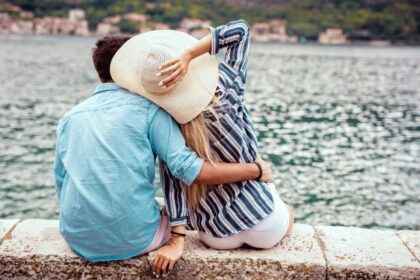 A couple sitting together on a rock wall, overlooking a lake with hills and buildings in the distance.