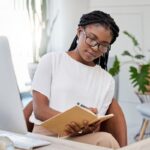 A woman working at her home office desk, using a computer and writing in a notebook.