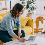 A woman working from home, sitting on a coach and using a laptop while her child plays in the background.