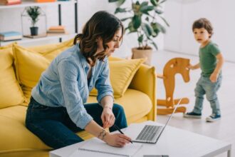A woman working from home, sitting on a coach and using a laptop while her child plays in the background.