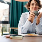 A woman working from her home office, sitting at a desk, writing in a notebook, and smiling while drinking coffee.