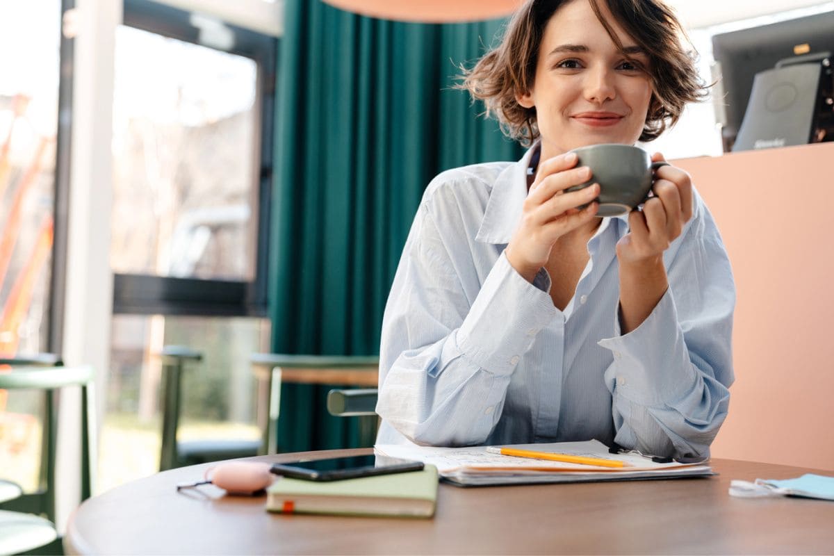 A woman working from her home office, sitting at a desk, writing in a notebook, and smiling while drinking coffee.