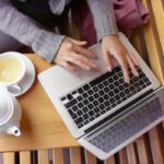 A closeup image of a woman's hands typing on a laptop while working remotely and drinking tea.