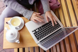 A closeup image of a woman's hands typing on a laptop while working remotely and drinking tea.