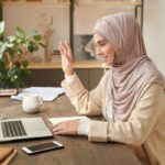 A woman using a laptop to take a video call from her home office, smiling and waving at the camera.