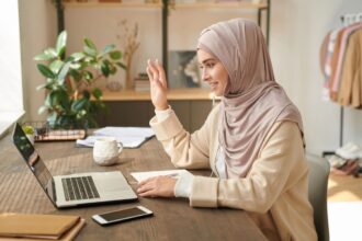 A woman using a laptop to take a video call from her home office, smiling and waving at the camera.