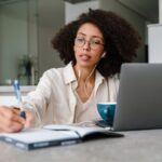 A woman working from home at her kitchen table, using a laptop and writing in a notebook.