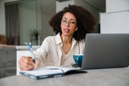 A woman working from home at her kitchen table, using a laptop and writing in a notebook.